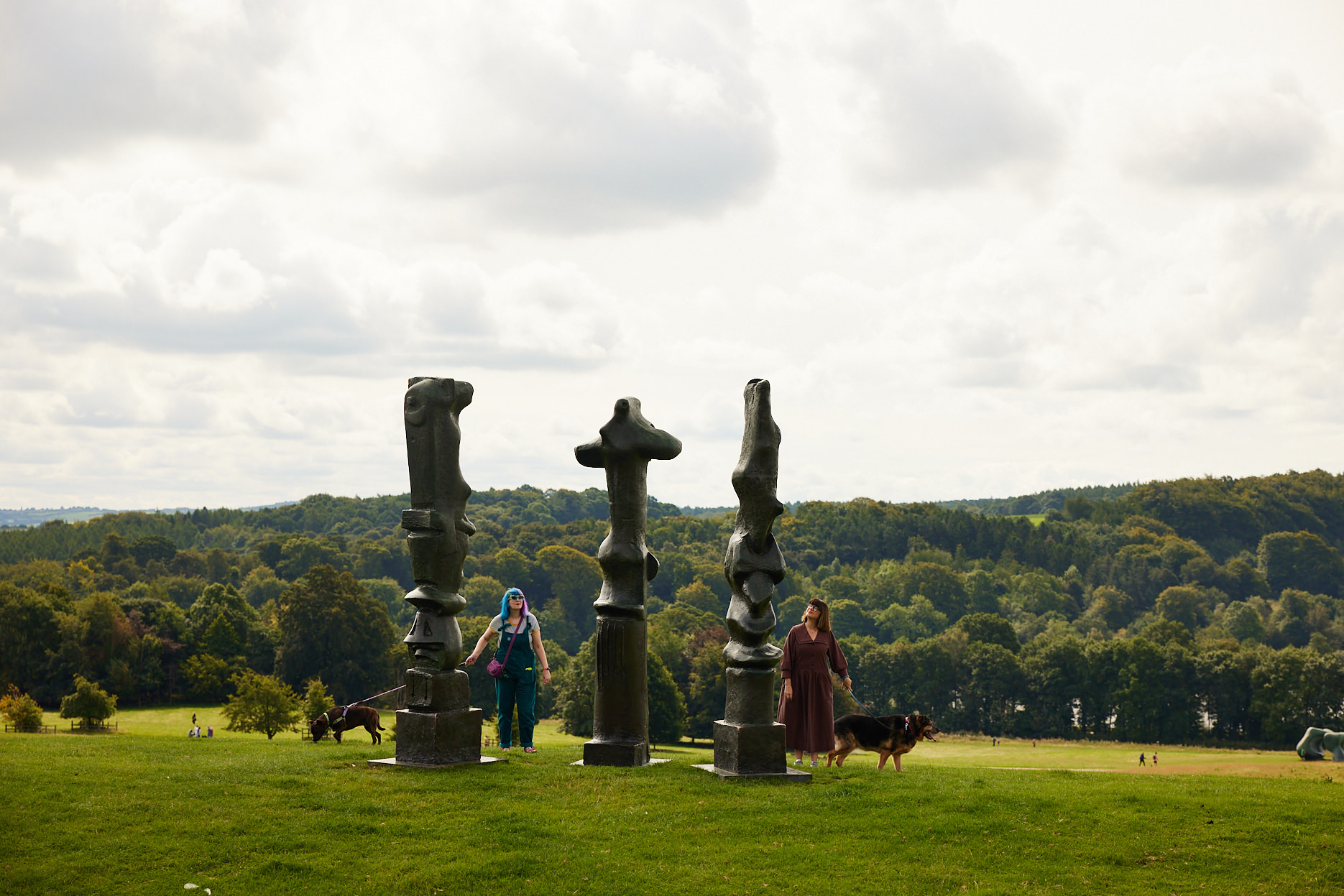 Dog walkers looking at three tall thin bronze sculptures looking out over the landscape