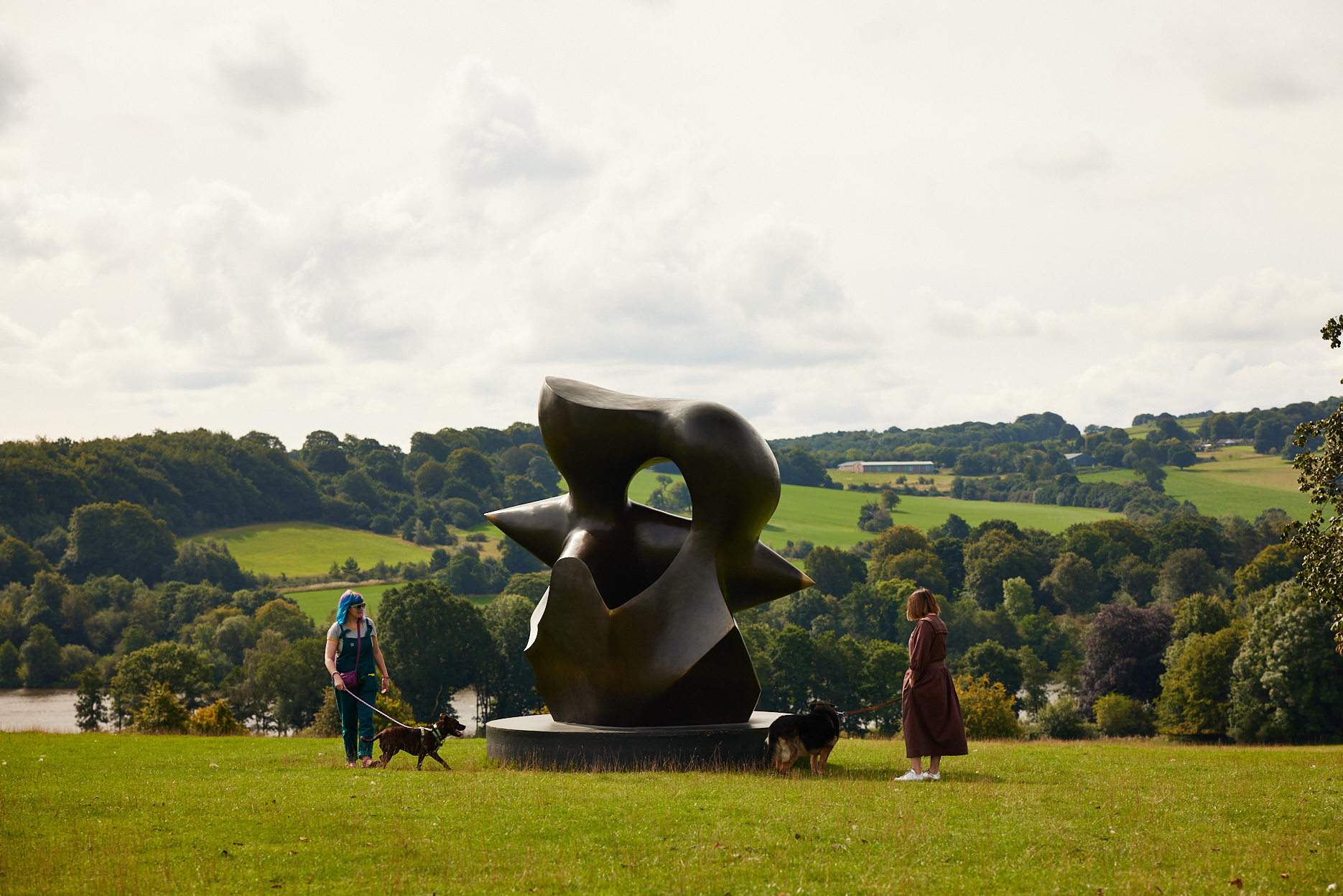 Two women with dogs on leads, looking at a large bronze Henry Moore sculpture outdoors