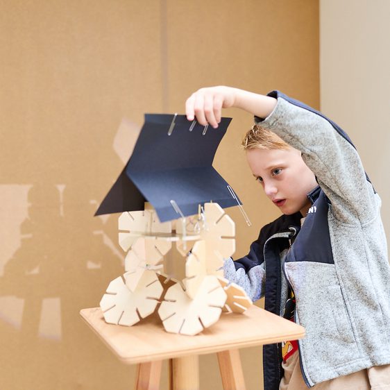 A young child with short hair is focused on assembling a wooden model on a small table. The model features wheels and a roof, and the child is carefully attaching parts with metal clips, showcasing creativity and concentration.