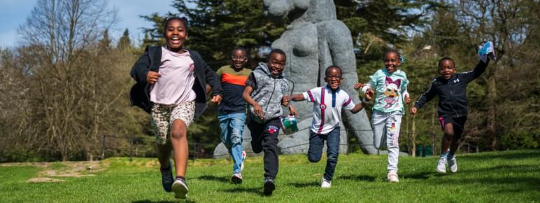 A group of six children joyfully run across a grassy field, smiling and laughing. In the background, a large sculpture of a rabbit stands among trees under a clear blue sky, creating a lively and playful atmosphere.