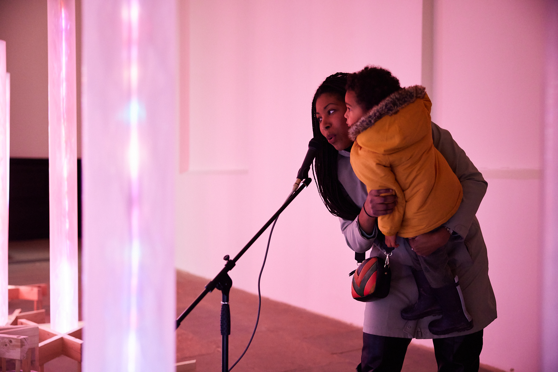 An adult holding a toddler, singing into a microphone surrounded by glowing columns.