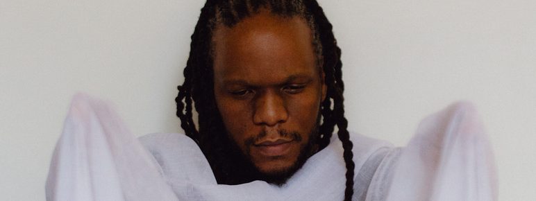 A man with braided hair is holding a sheer, light-coloured fabric in front of him. He appears contemplative, with his eyes closed, and is dressed in a simple, flowing garment. The background is plain and neutral, enhancing the focus on him.