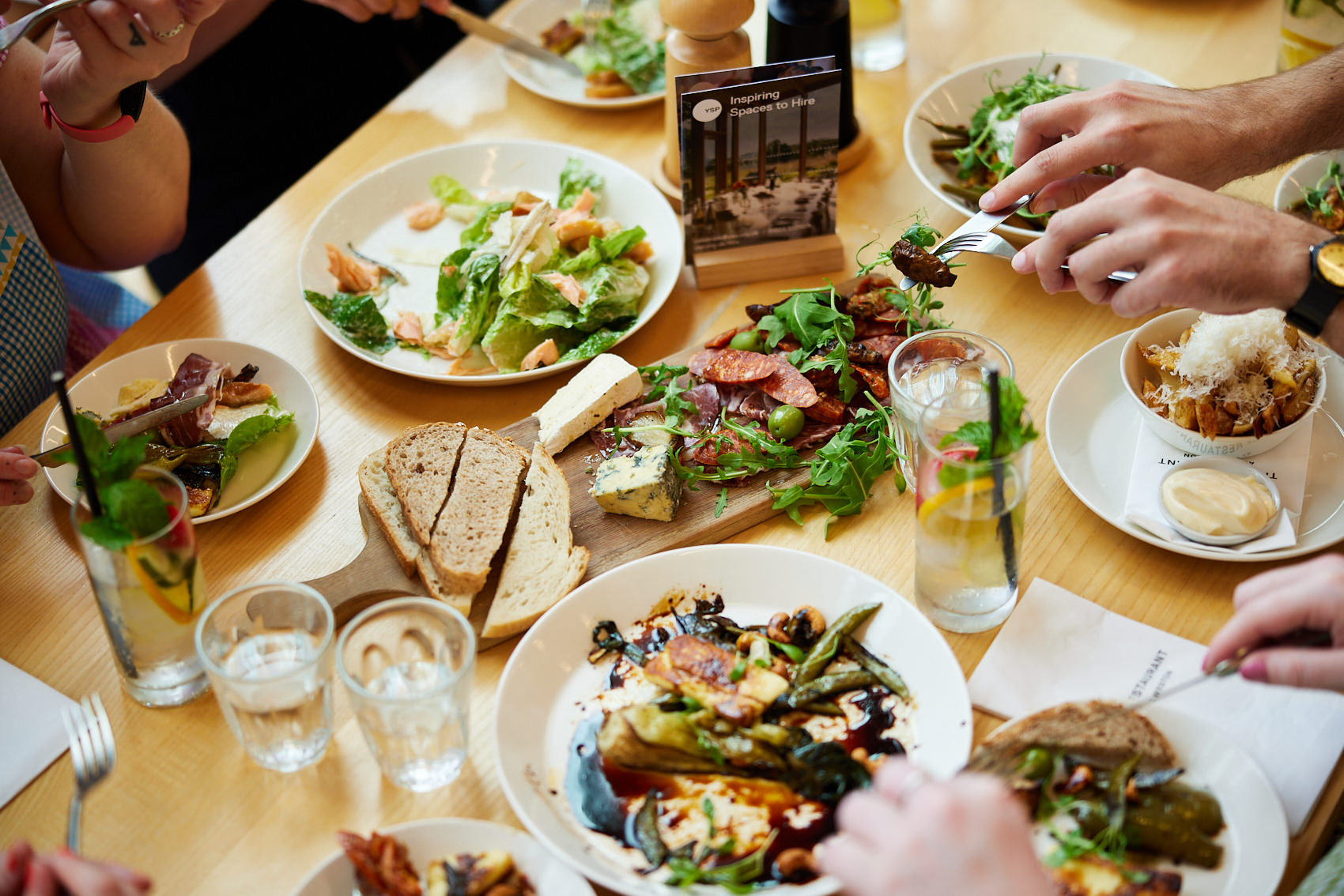 A table filled with small plates of food while hands holding forks reach for the food.