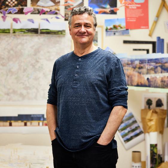A man with short grey hair standing with his hands in his pockets in an art studio. Behind him are drawings in progress pinned to the walls.