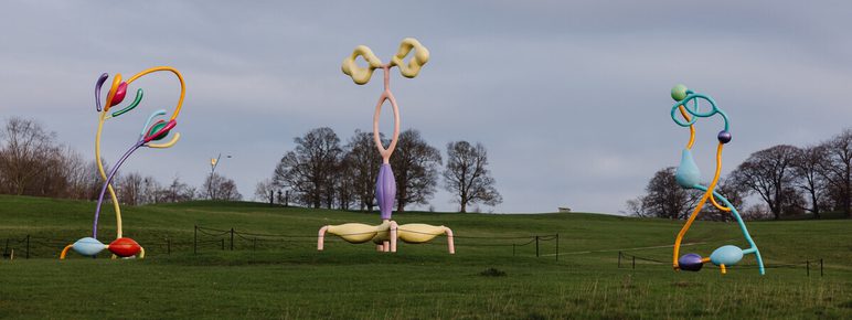 Three colourful abstract sculptures displayed outdoors in parkland, with trees in the distance.