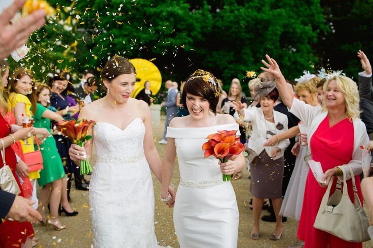 A pair of brides in white wedding dresses, walk between two rows of friends and family throwing petal confetti.