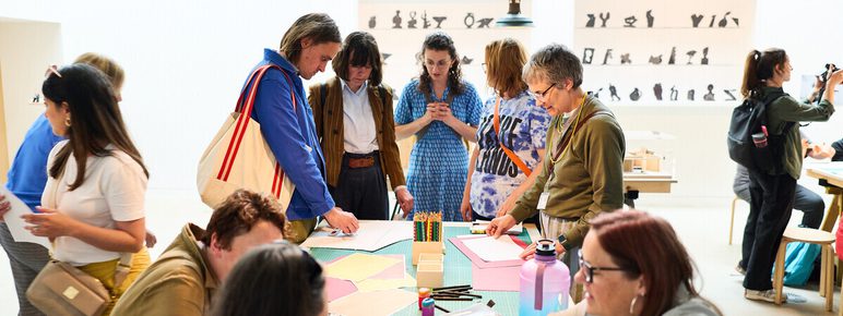 A group of people engage in a creative workshop in a bright, modern space. Some are gathered around a table covered with craft materials, while others are focused on their phones or chatting. The walls feature artistic silhouettes.