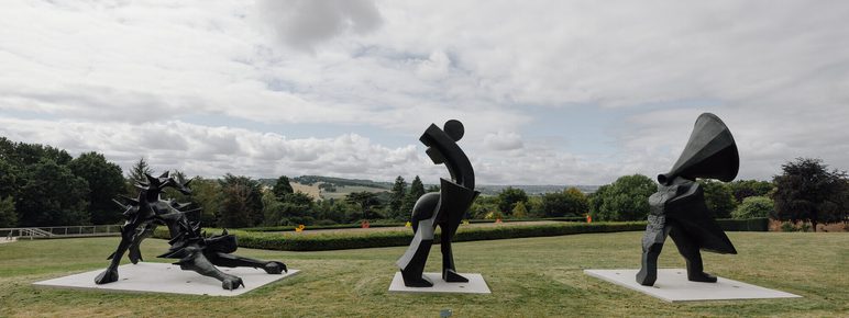 Three abstract sculptures are displayed on a grassy area, set against a cloudy sky. The first sculpture features dynamic, twisted forms; the second is a stylised figure in motion; the third depicts a figure with a large, flowing cape. Lush greenery surrounds them.