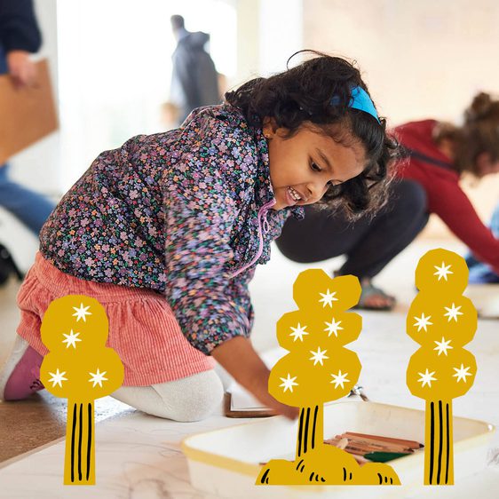 A young girl is kneeling on the floor, focused on drawing with crayons on a large sheet of paper. In the background, other children and adults are engaged in various art activities in a bright, spacious room. Illustrations of trees are overlaid onto the image.