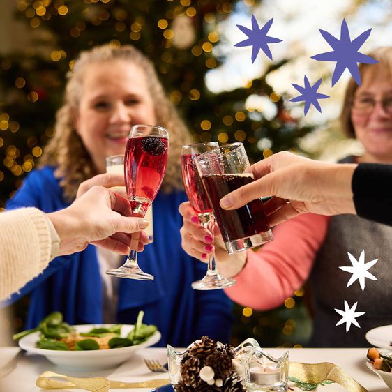 A festive gathering featuring three people toasting with drinks at a table adorned with food. In the background, a decorated Christmas tree glimmers with lights.