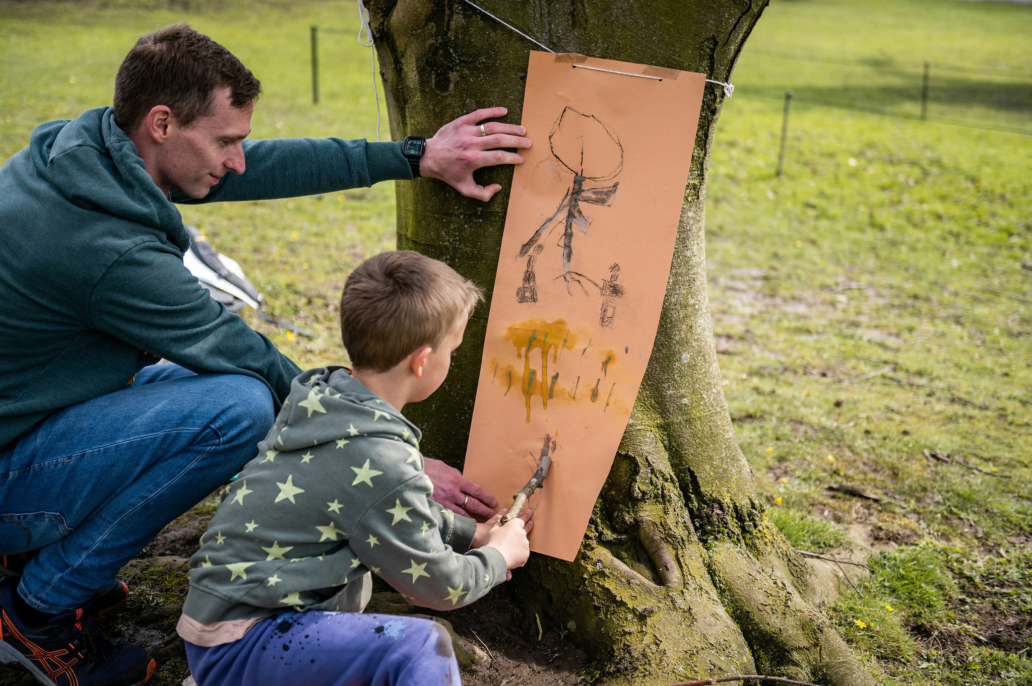 An adult and a child drawing together onto a large piece of paper leaning against a tree outdoors.