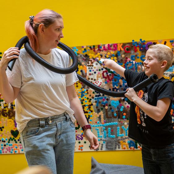 An adult and a child with limb difference, playing with black tubes. Behind them is a mirror covered in colourful circular stickers.