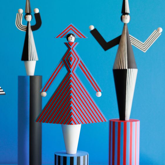 Three whimsical figurines stand on cylindrical pedestals against a bright blue background. The central figure, dressed in a red and black striped dress with a matching hat, is flanked by two others in contrasting patterns, creating a playful, artistic display.