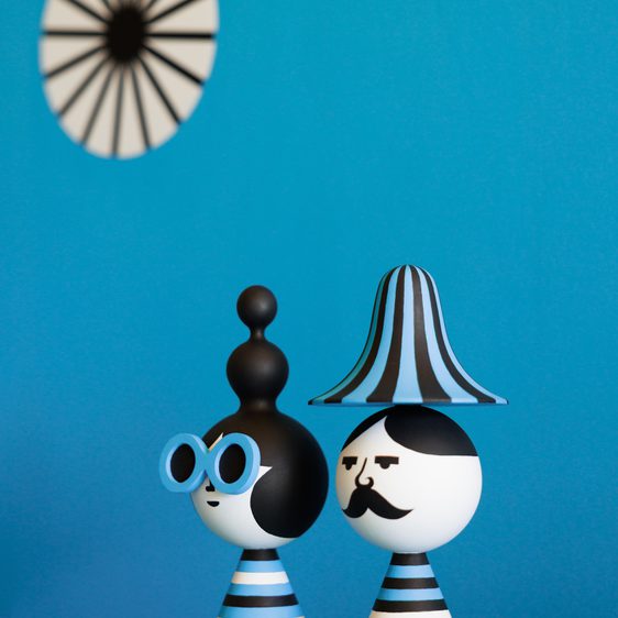 Two whimsical figurines stand on a striped table, one with oversized blue glasses and a round black hat, the other with a striped hat and a painted moustache. The background features a blue wall and a circular black-and-white design.
