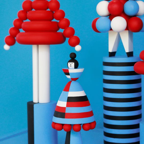 A vibrant display of abstract wooden figures in red, white, and blue. The figures include a striped column, a round-topped structure, and a stylised woman in a dress with bold patterns, all set against a bright blue background.
