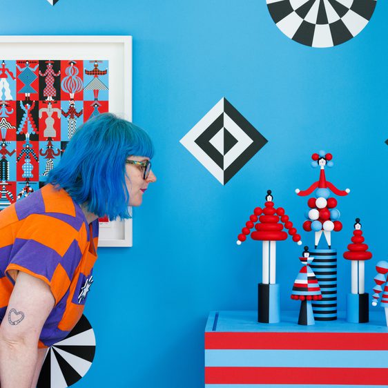 A woman with blue hair leans towards a display of colourful, abstract sculptures on a table. The background features a vibrant blue wall adorned with geometric patterns in red, black, and white, alongside a framed artwork.