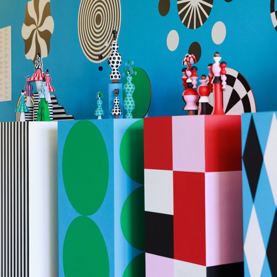 A vibrant display featuring geometric shapes in bold colours, including green circles, red and pink blocks, and black and white patterns. The background is a bright blue adorned with swirling designs and playful motifs, creating a dynamic visual effect.