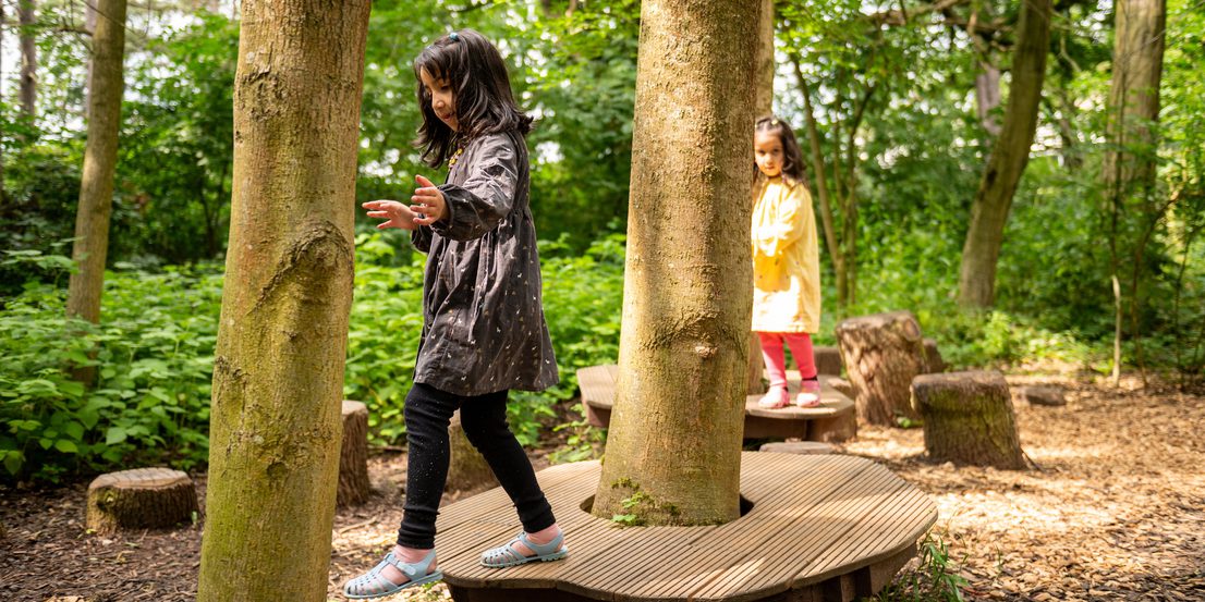 Two children jumping between wooden platforms in a nature play area.