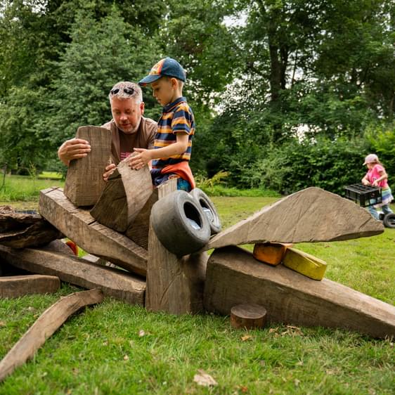 An adult and child building a sculpture out of pieces of wood in an outdoor setting.