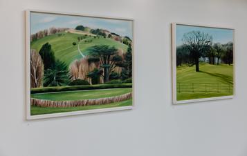 Two landscape paintings depicting tress and hills displayed on a gallery wall.
