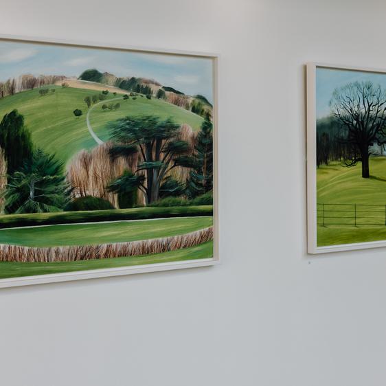 Two landscape paintings depicting tress and hills displayed on a gallery wall.