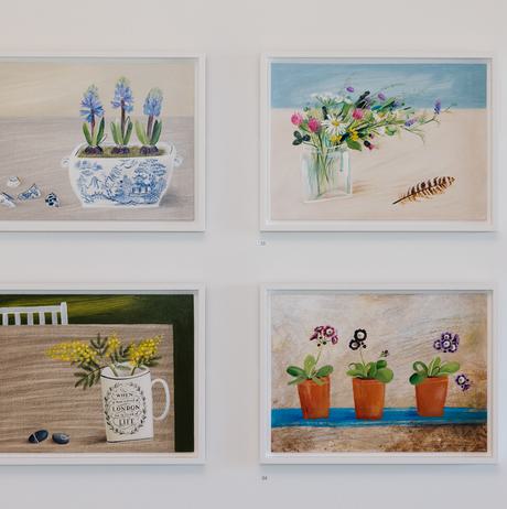 Four framed paintings of flowers in still life scenes, displayed in a grid on a gallery wall.