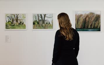 A woman in a black dress looking at three frames landscape paintings displayed on a gallery wall.