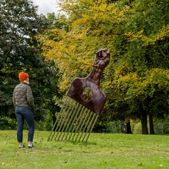 A woman wearing a coat and wooly hat looking at a sculpture of an oversized afro pick with a black power as a handle, displayed outdoors.