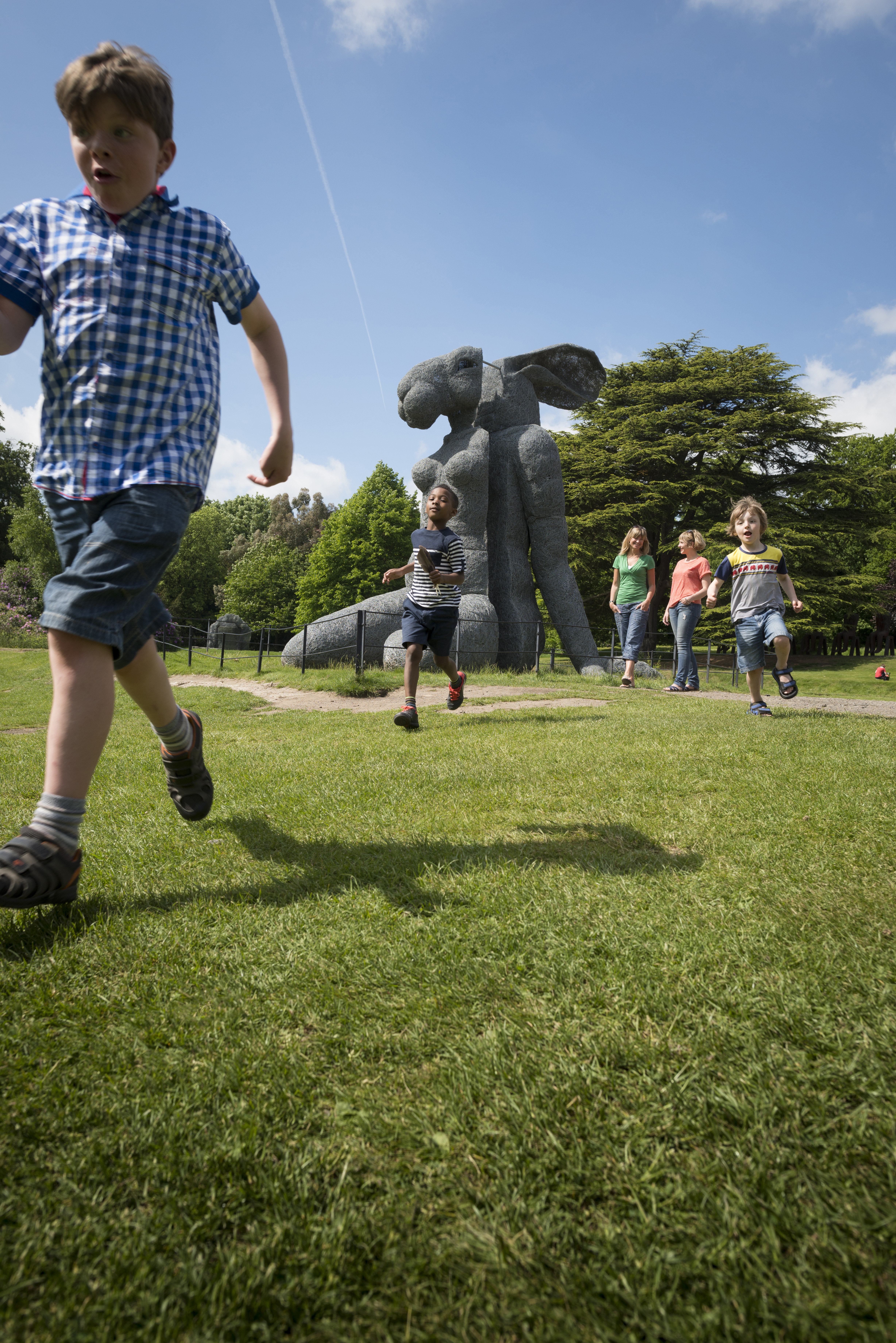 A group of children running in front of a large sculpture of a female body with a rabbit head.