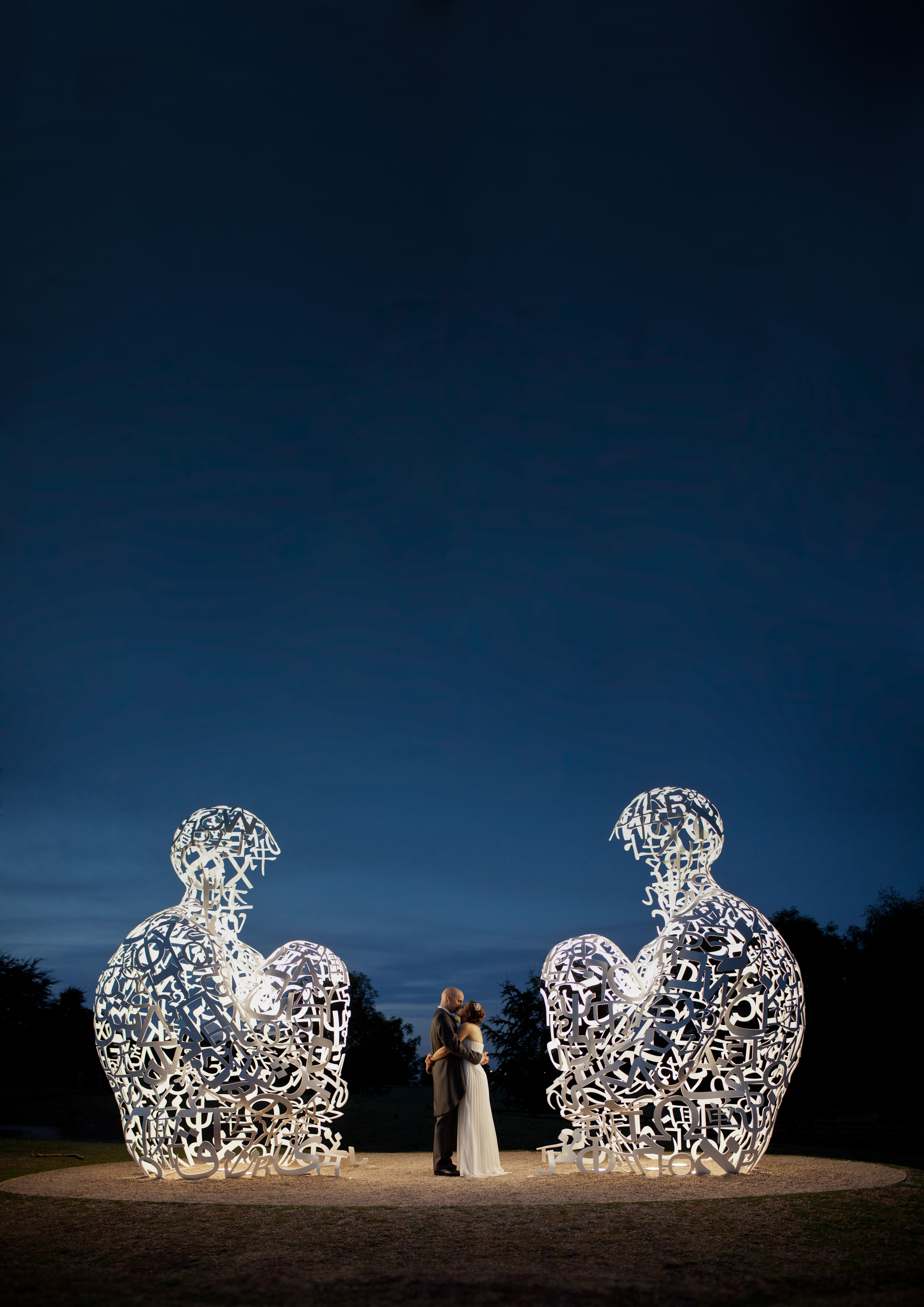 A bride and groom standing between two sculptures of sitting human figures at night.