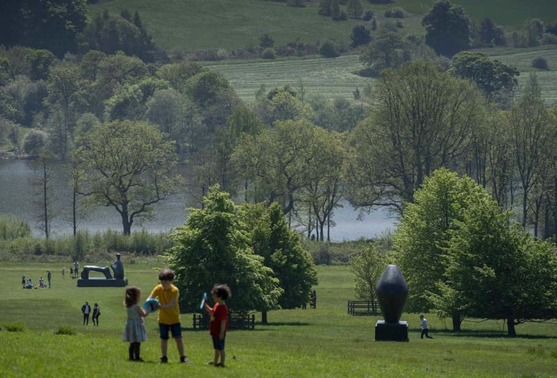 Children playing outdoors on a sunny day. A rolling landscape with trees and sculptures is behind them.