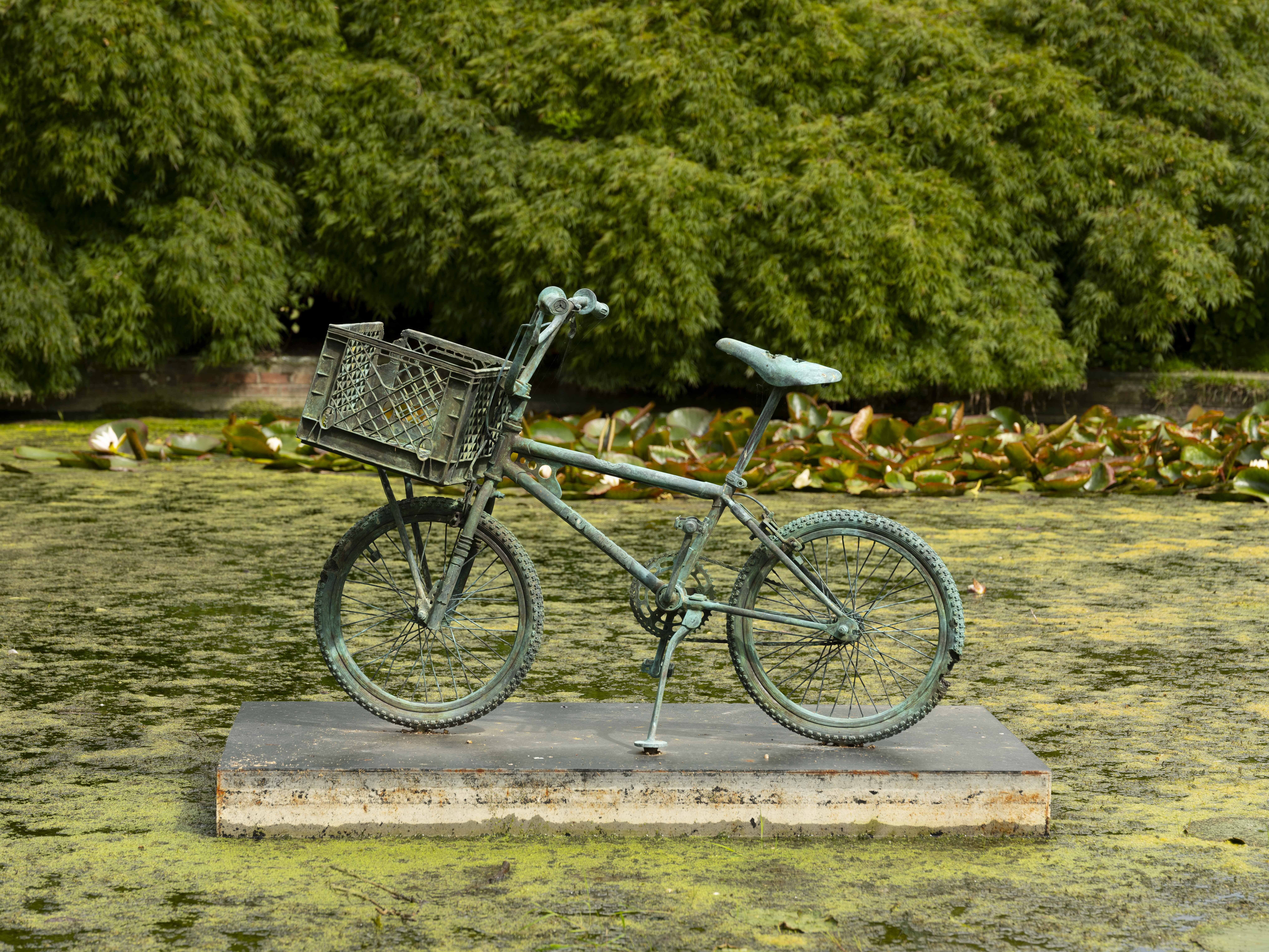 A bronze bicycle mounted on a plinth in the centre of a pond, with bushes and trees in the background.