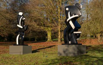 Two black and white abstract sculptures displayed on plinths outdoors, with trees behind them.