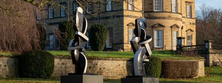 Two black and white abstract sculptures displayed on plinths outdoors. A historic manor house is behind them.