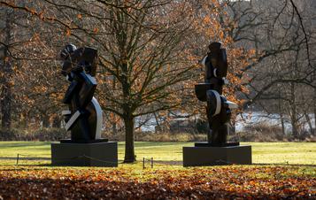 Two black and white abstract sculptures displayed on plinths outdoors, with trees behind them.