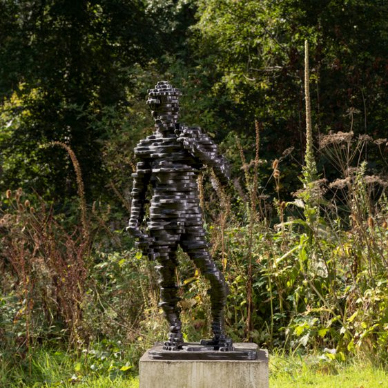 A sculpture of a human figure made from layers of bronze, displayed on a concrete plinth outdoors with foliage behind it.