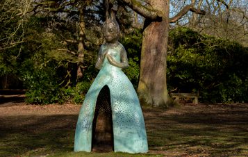 A sculpture of a figure with a rabbit head and a large blue skirt with a hollow opening in the front, displayed outdoors.