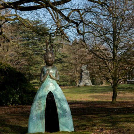 A sculpture of a figure with a rabbit head and a large blue skirt with a hollow opening in the front, displayed outdoors.