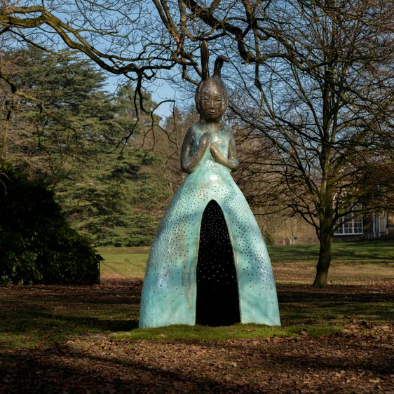 A sculpture of a figure with a rabbit head and a large blue skirt with a hollow opening in the front, displayed outdoors.
