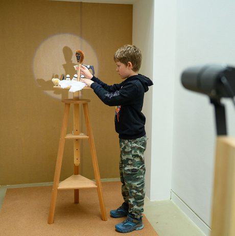 A young boy stands at a wooden stool, focused on a small model illuminated by a circle of light. The scene is set in a minimalist room with a plain wall, and a directional light is visible in the foreground.