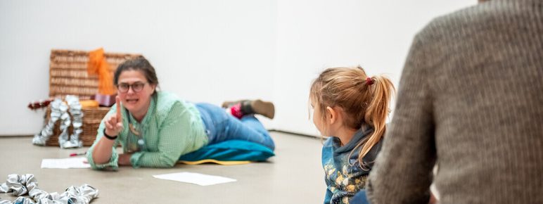 A storyteller lies on a gallery floor in front of a box filled with colourful objects. A child and parent watch.