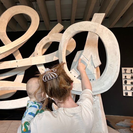 A woman holds a child as they admire a large, abstract sculpture made of curved, white forms. The sculpture is set against a dark background, and the woman points towards a specific part of the artwork, engaging the child’s curiosity.