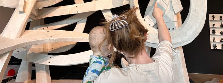 A woman holds a child as they admire a large, abstract sculpture made of curved, white forms. The sculpture is set against a dark background, and the woman points towards a specific part of the artwork, engaging the child’s curiosity.
