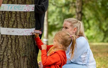 A woman and child drawing on thin strips of paper fastened around the trunk of a tree.