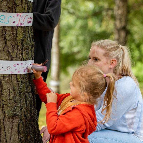 A woman and child drawing on thin strips of paper fastened around the trunk of a tree.