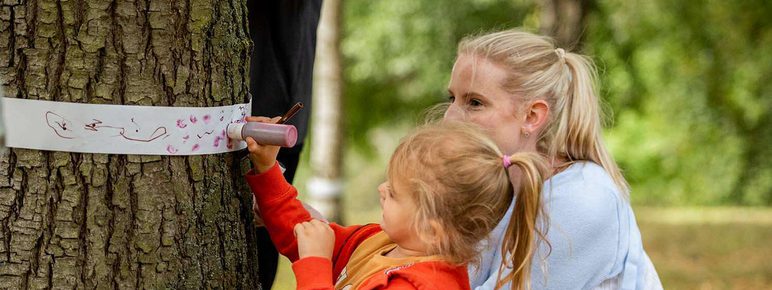 A woman and child drawing on thin strips of paper fastened around the trunk of a tree.