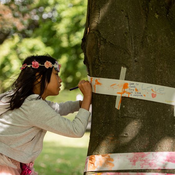 A child drawing on to strips of paper which have been wrapped around the trunk of a tree.