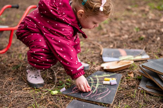 A young girl in a red polka-dot snowsuit kneels on the ground, picking up chalk to draw on a slate board. Surrounding her are several other slate boards and a wooden bowl, set in a natural outdoor environment.