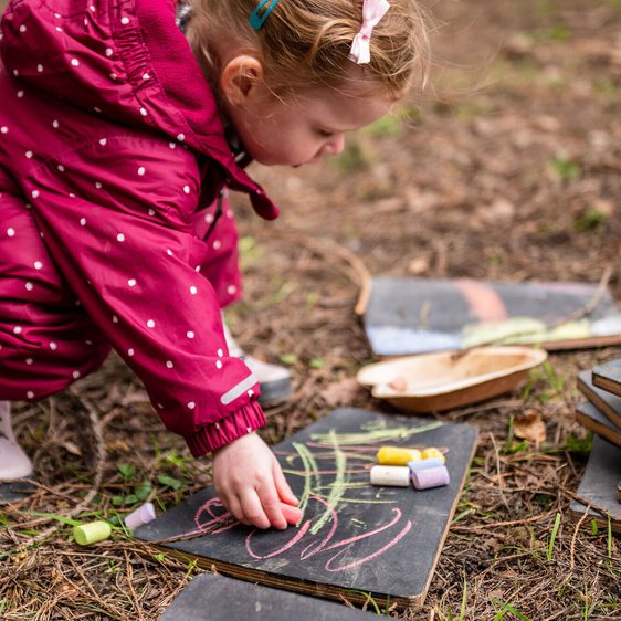 A young girl in a red polka-dot snowsuit kneels on the ground, picking up chalk to draw on a slate board. Surrounding her are several other slate boards and a wooden bowl, set in a natural outdoor environment.