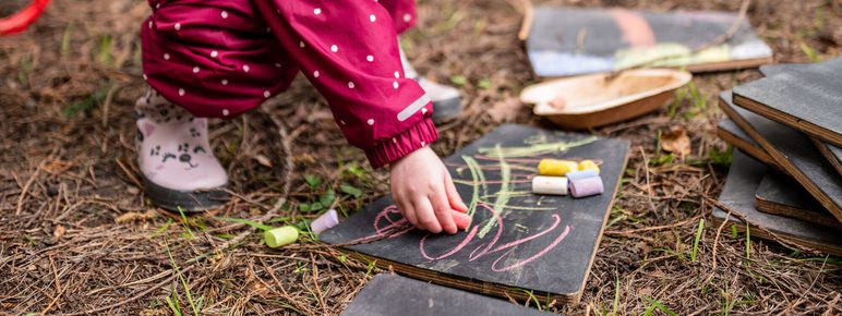 A young girl in a red polka-dot snowsuit kneels on the ground, picking up chalk to draw on a slate board. Surrounding her are several other slate boards and a wooden bowl, set in a natural outdoor environment.